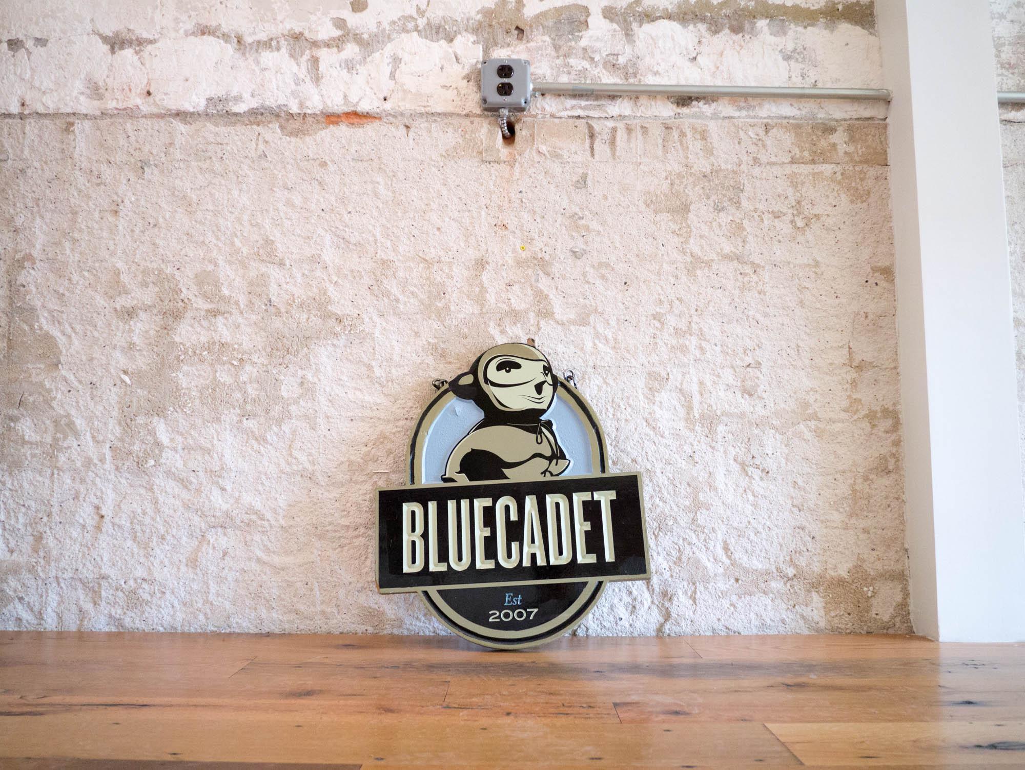 A metal sign with the words Bluecadet, est. 2007. Behind the words is an illustrated character in a heroic pose.