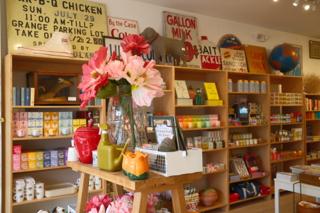 Paper flowers in a vase in front of shelves with books, candles, and other goods.