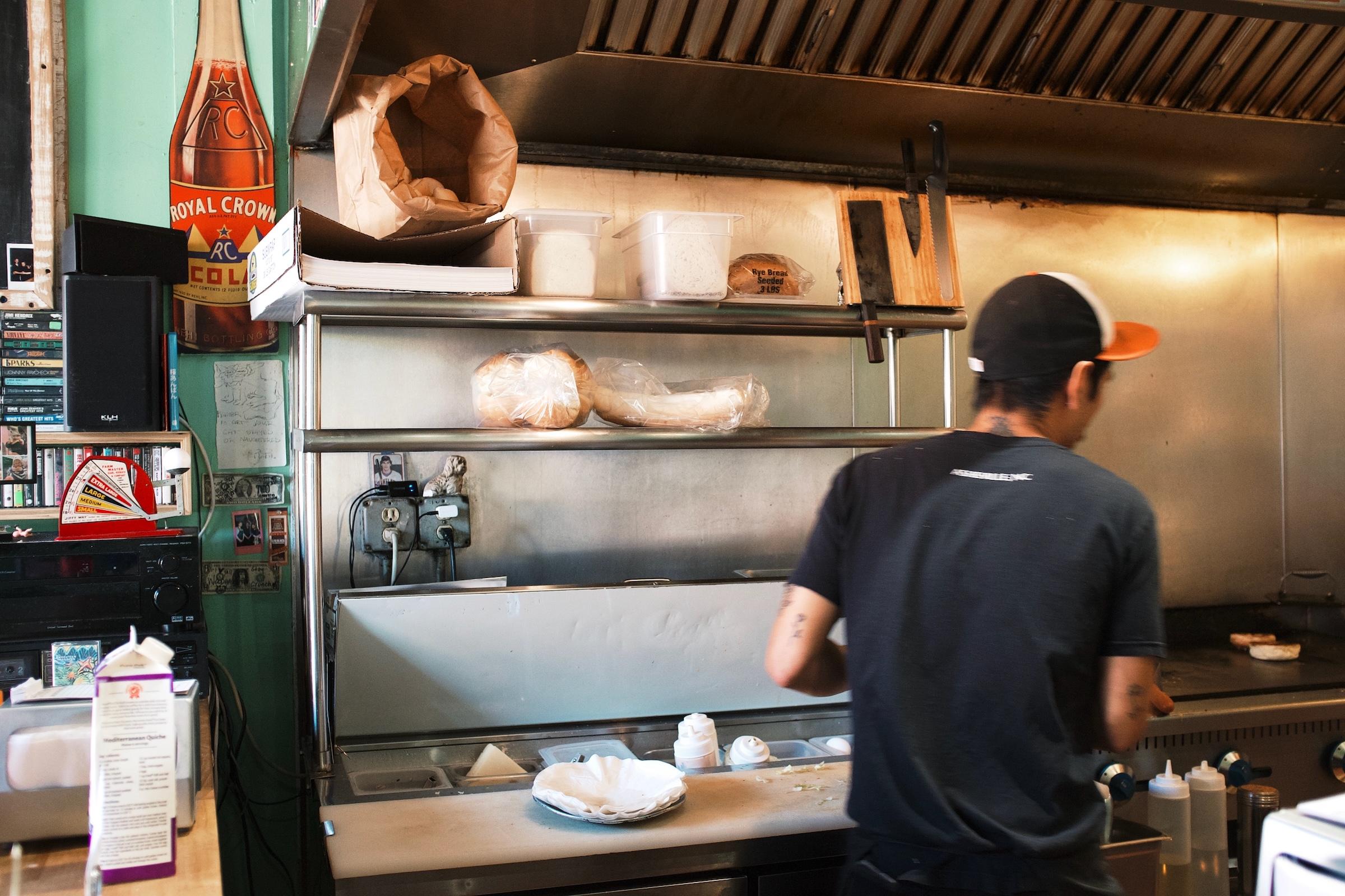 A cook wearing an orange-brimmed cap preps a sandwich behind the counter at a restaurant.