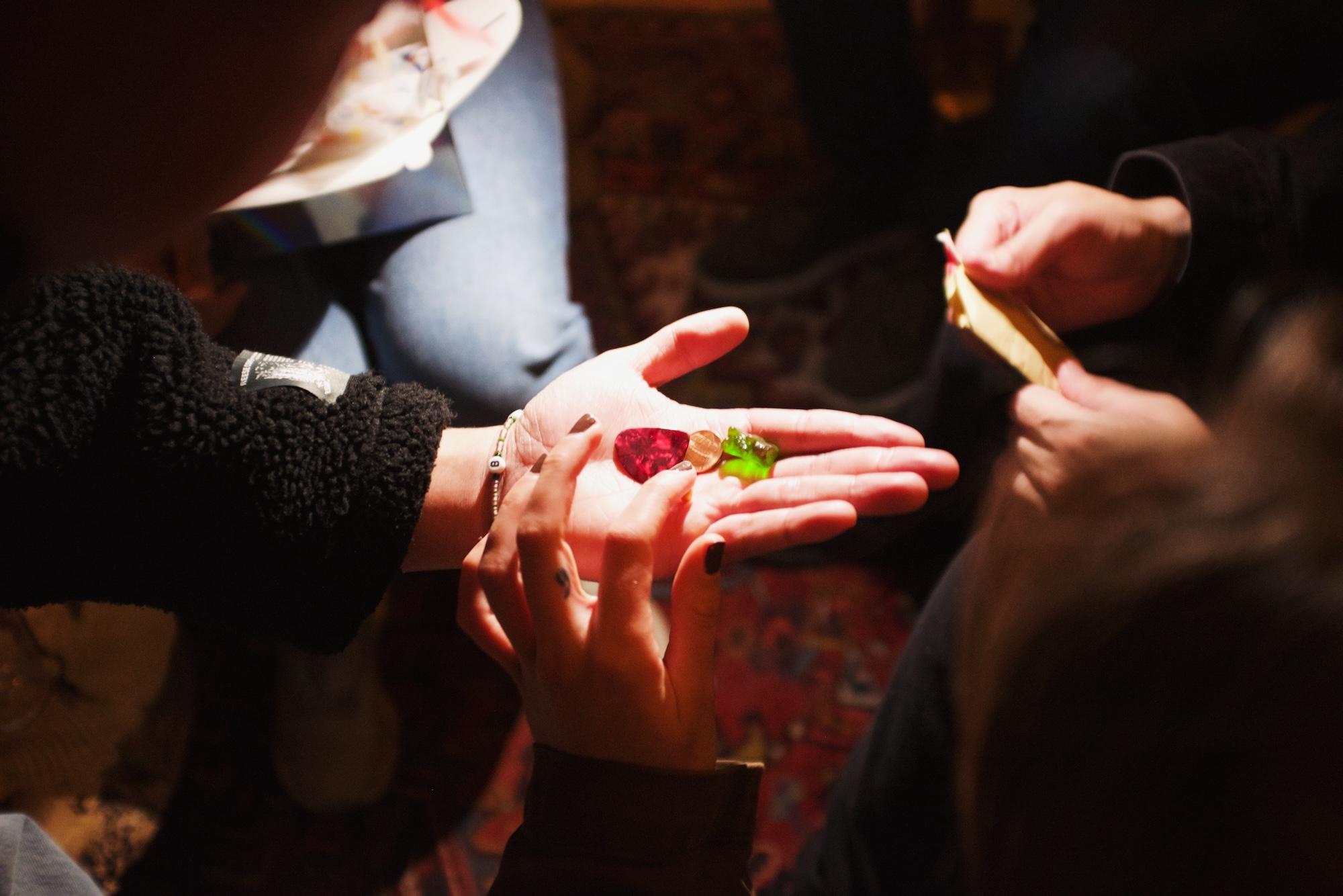 Multiple people huddle over a hand holding a guitar pick, a penny, and a gummy bear.