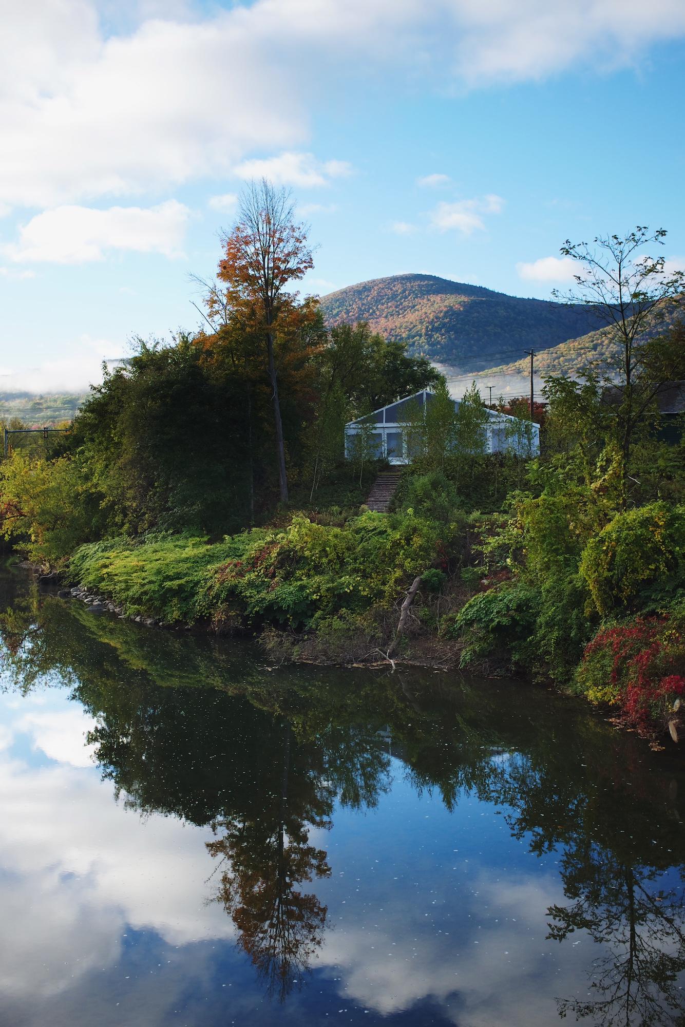 A riverbank with a mountain in the distance the trees and sky are reflected in the river.