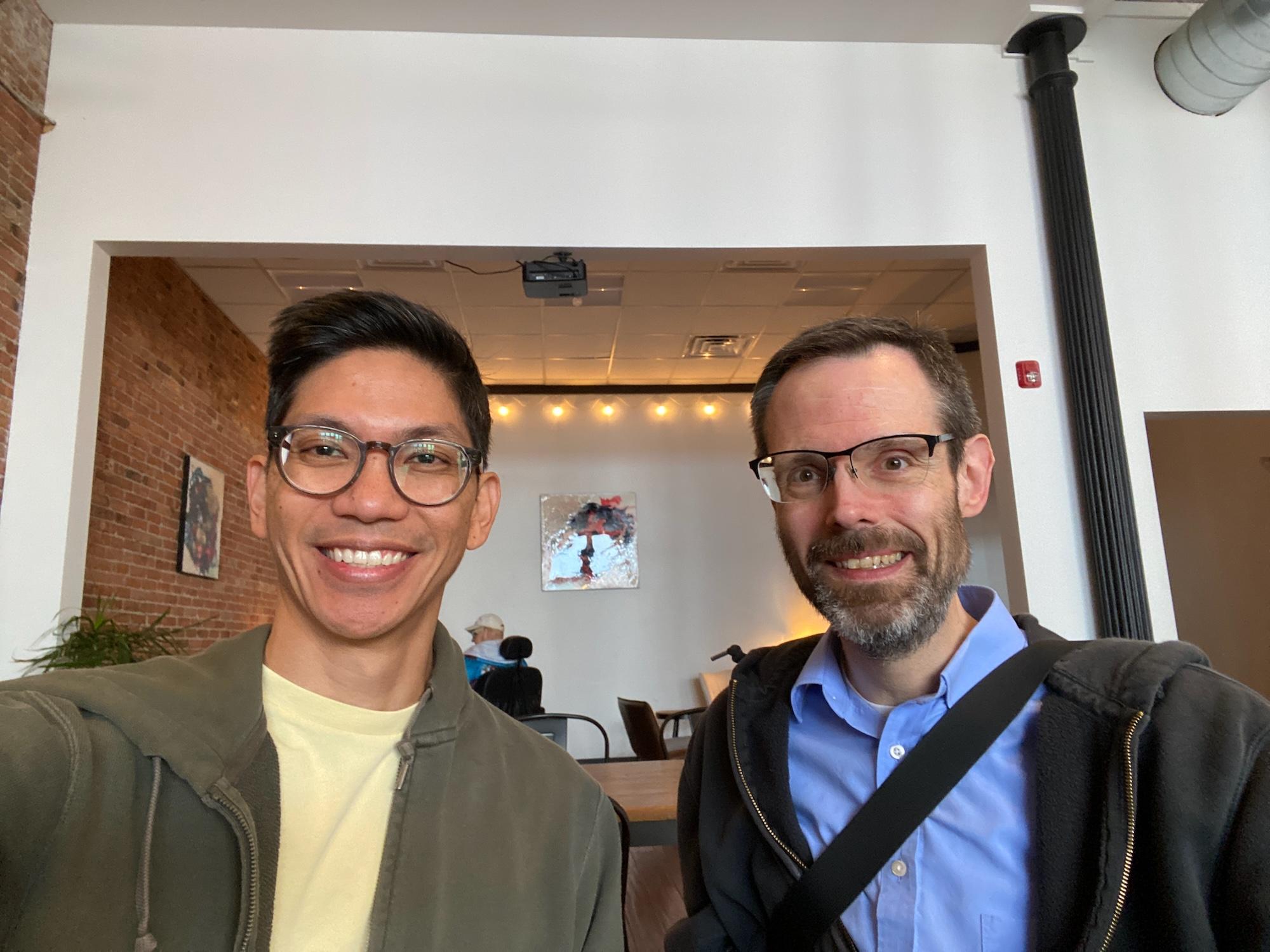 Two men smile in a coffee shop selfie.