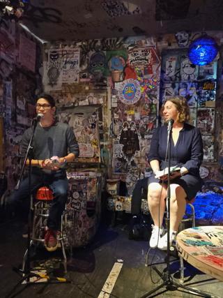 A man and a woman sit side-by-side on bar stools in a makeshift performance space. Both of them have microphones and the woman is listening as the man asks her a question.
