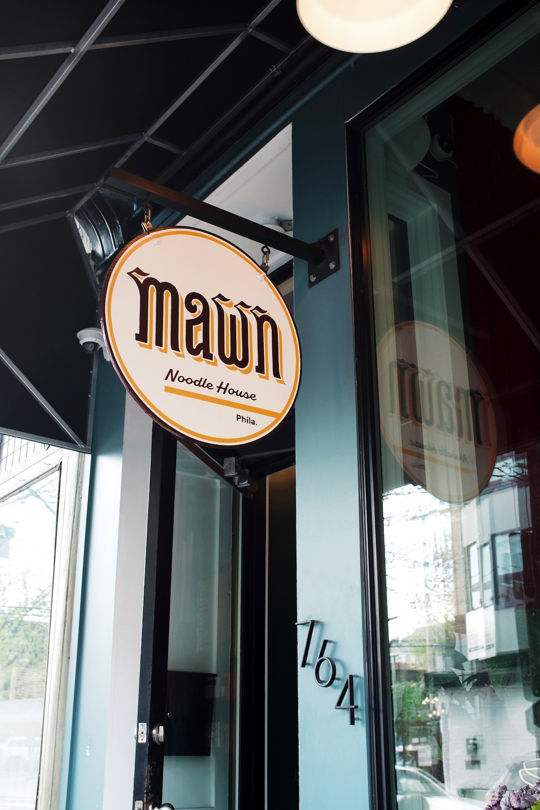 A round sign for Mawn Noodle House in Philadelphia. The sign hangs from a post and is reflected in the tall window at the front of the restaurant