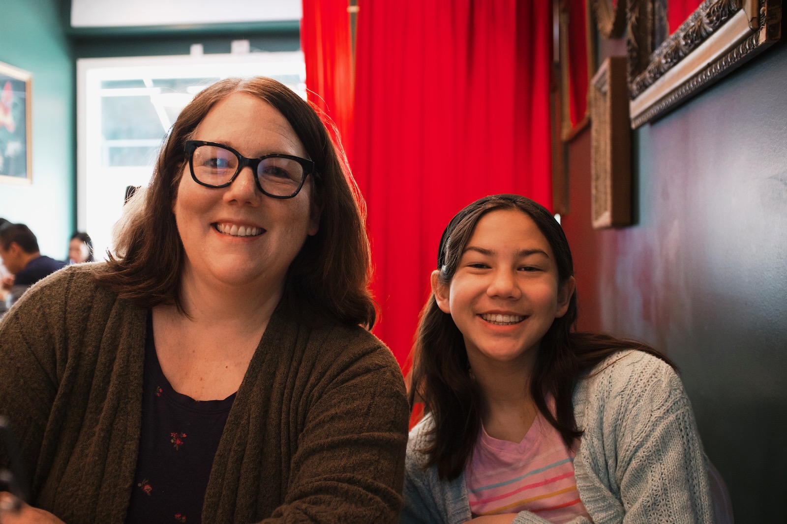 A woman and a young girl sitting side by side in a restaurant, smiling. The woman wears glasses and wears a green cardigan. The girl has a headband and a pink shirt with a grey cardigan over it. Behind them in the near distance is a bright red curtain leading to the entrance of the restaurant.