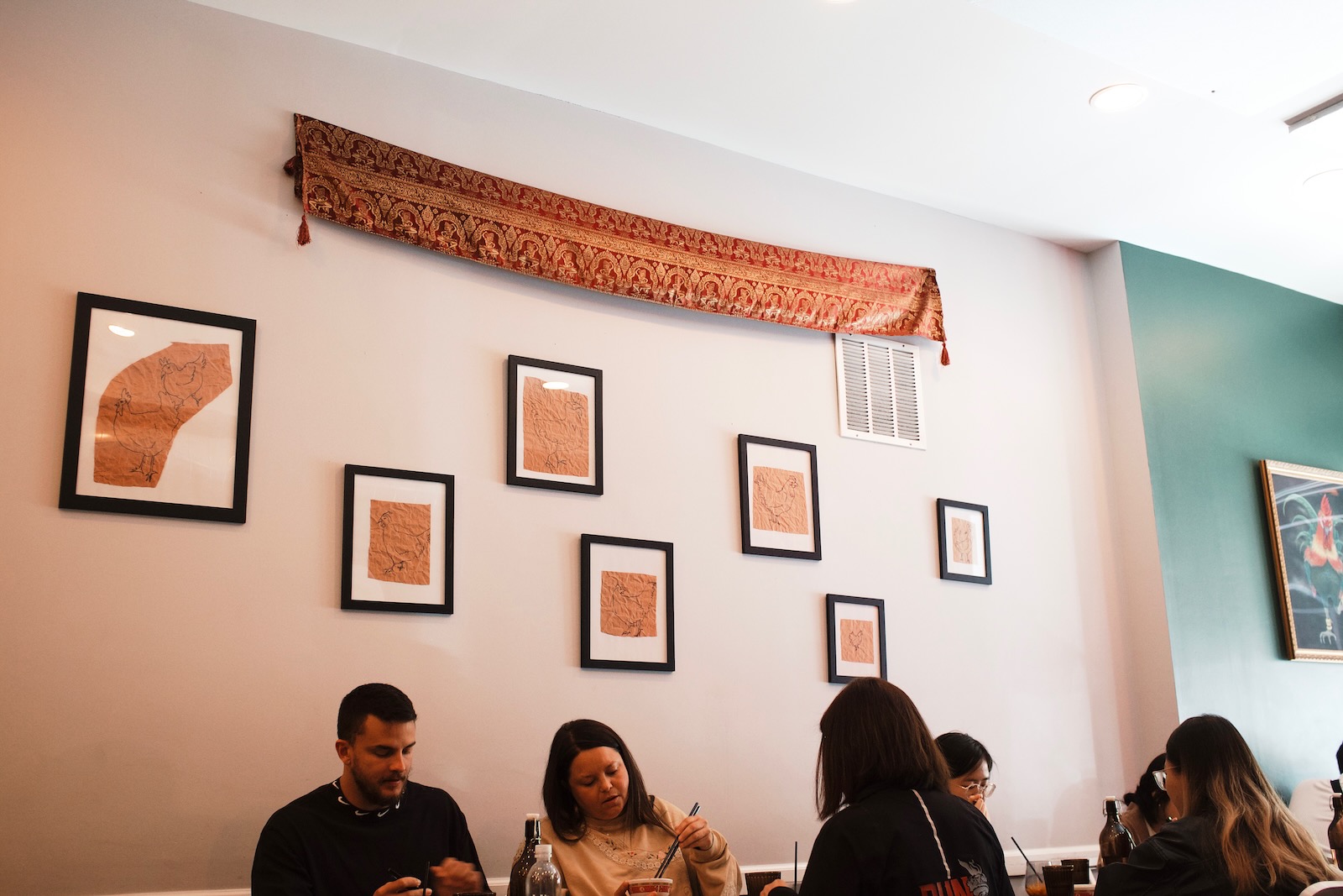 Diners share a meal. In the wall behind them is an arrangement of several framed prints of illustrated chickens, and an elaborate printed cloth stretched across the top of several of the frames.