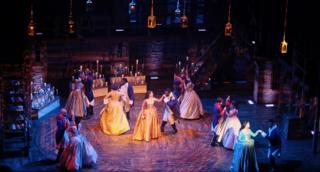 A party scene from “Hamilton” featuring women in colorful gowns and men in uniform.