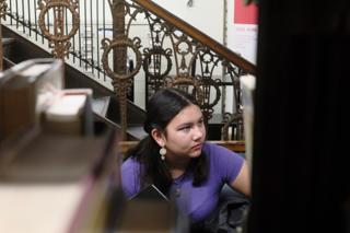 A young woman listens to a college tour. She is visible between the books in a library shelf.