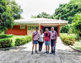 A family of four stands in the driveway in front of a low-slung red house.