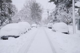View down a suburban street, with parked cars on either side covered in snow.