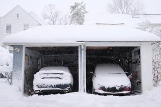 Side-by-side garages with no doors. The cars parked are covered lightly in snow. Snow falls gently in the foreground.