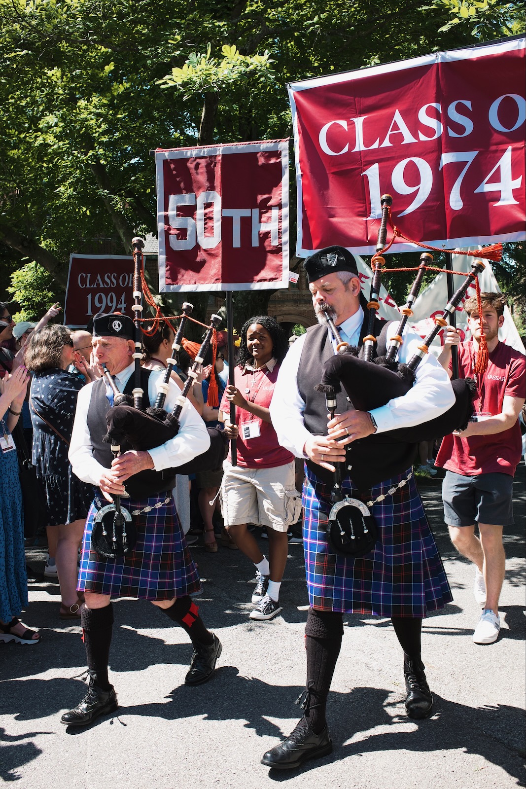 Two men wearing kilts play bagpipes in front of students holding up a banner reading “Class of 1974”.
