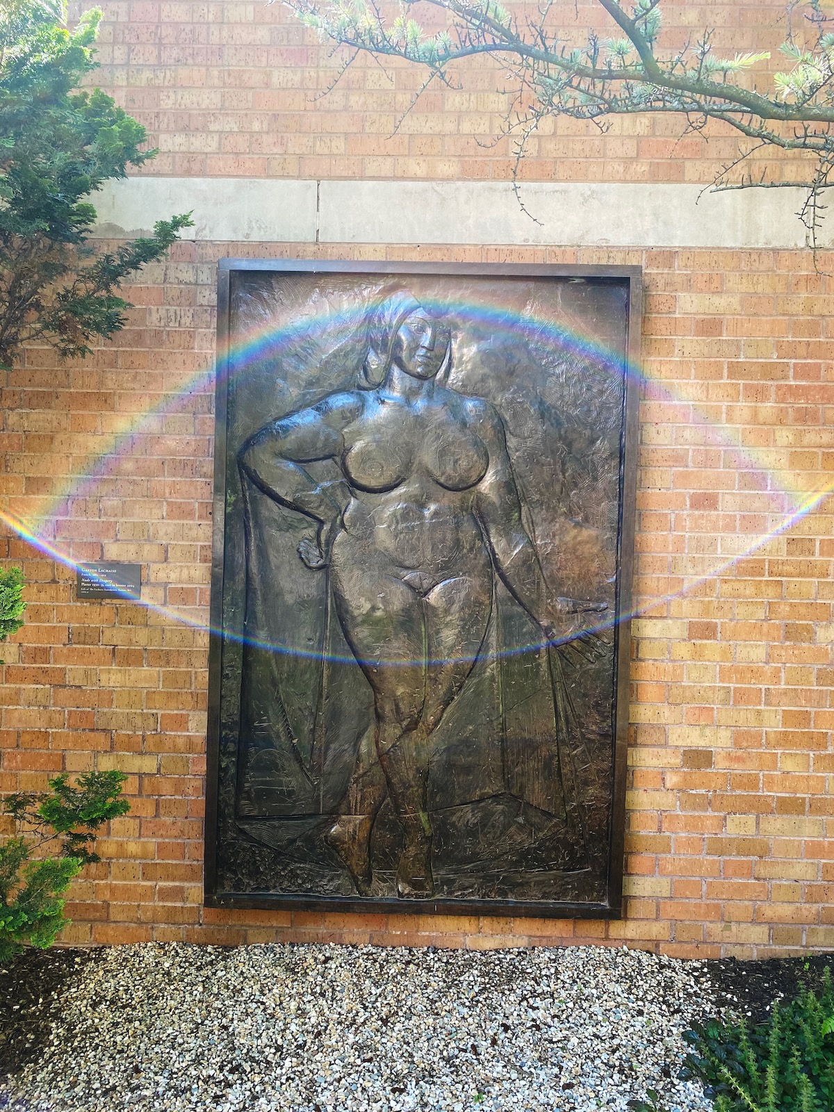 A bronze sculpture of a nude woman with cloth draped about her head and shoulders, mounted on a brick wall. Light streaming over the wall is creating a lens flare shaped like an eye.