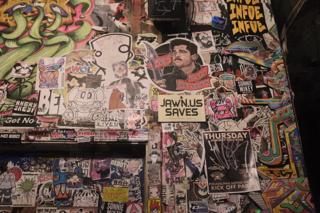 Close up on a section of a wall, covered with stickers. One sticker reads “Jawn.us saves”.