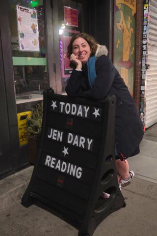 A woman poses, leaning, on an outdoor bar sign reading “Today Jen Dary Reading"