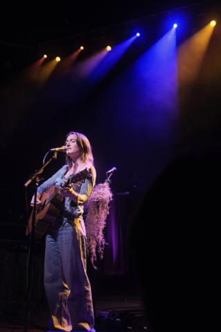 A woman sings while playing guiter, lit from behind by blue and yellow spots.