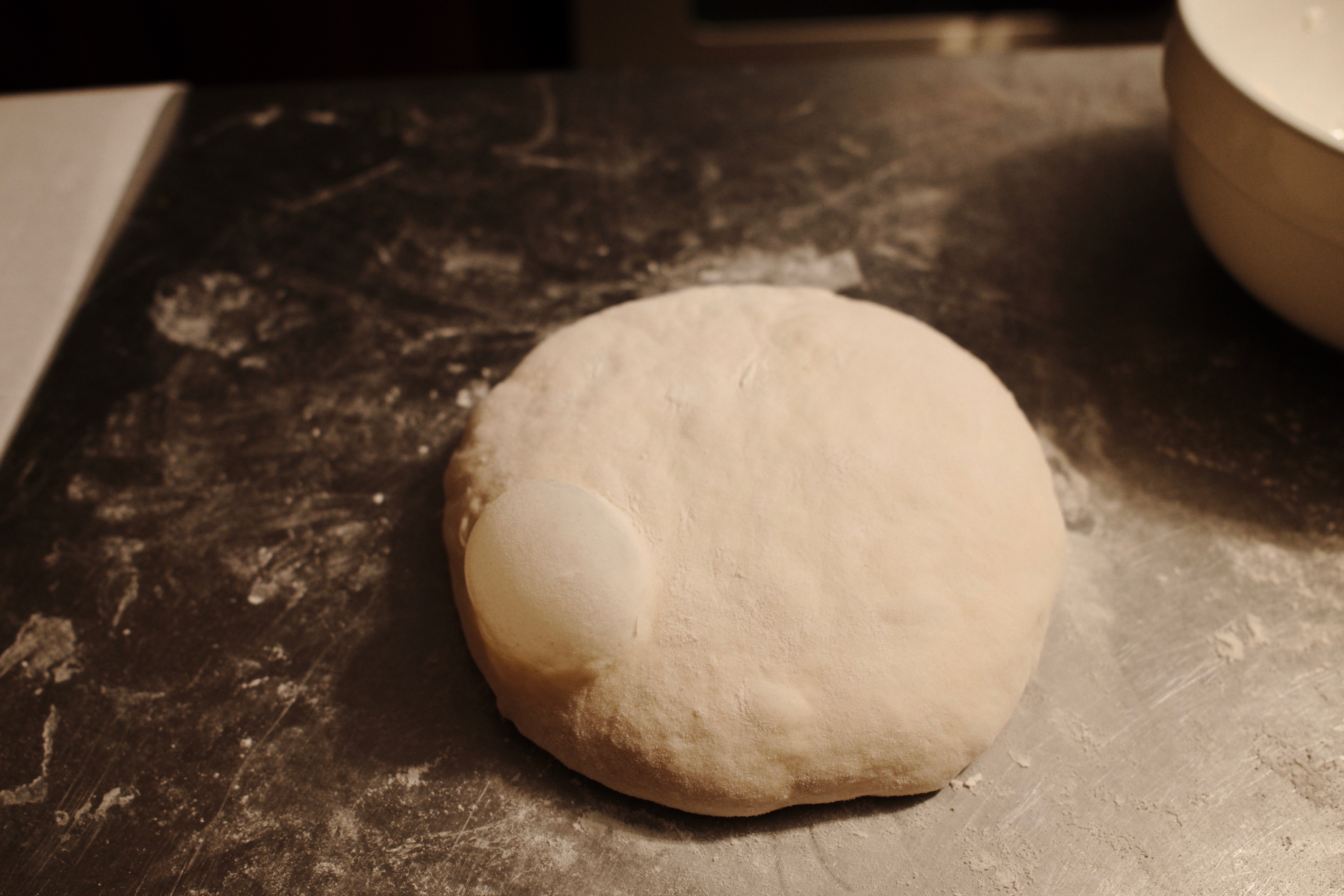 Pizza dough round resting atop a countertop. A big bubble forms on the edge.